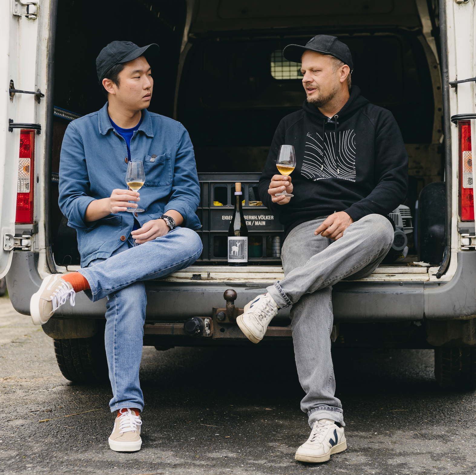 Two men sitting in the back of a van, holding wine glasses.